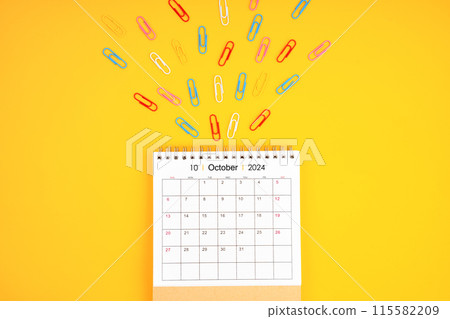 October 2024 desk calendar and colorful metal paperclips on yellow background. 115582209
