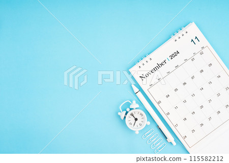 November 2024, Monthly desk calendar for 2024 year and alarm clock with paper clips on blue color. November 2024, Monthly desk calendar for 2024 year and alarm clock with paper clips on blue color. 115582212