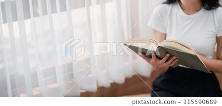 A woman is reading a book in a room with white curtains. The curtains are drawn, and the room is dimly lit. The woman is sitting on a chair, and she is holding the book in her hands 115590689