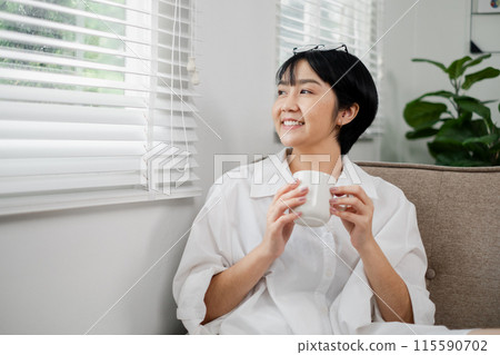 Young woman enjoying a cup of coffee while looking out the window in a bright, modern living room with natural light and green plants. 115590702