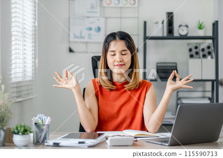 A woman in an orange shirt is sitting at a desk with a laptop and a book. She is practicing yoga and she is in a calm and focused state 115590713