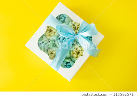 White gift box with pastel blue sweets inside, tied with a blue ribbon on a yellow background. White gift box with pastel blue sweets inside, tied with a blue ribbon on a yellow background. 115592571