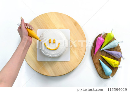Closeup woman hand is icing a smile face on a white cake. Colorful icing bags are placed aside. Top view Closeup woman hand is icing a smile face on a white cake. Colorful icing bags are placed aside. Top view 115594029