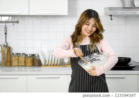 A happy woman in an apron, mixing batter in a clear glass bowl with a wooden spatula. Standing in a clean and modern kitchen A happy woman in an apron, mixing batter in a clear glass bowl with a wooden spatula. Standing in a clean and modern kitchen 115594397