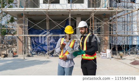 Two construction workers checking work schedule on tablet computer. Wear hardhat and safety vest, stands by metal scaffolding at a building site. Two construction workers checking work schedule on tablet computer. Wear hardhat and safety vest, stands by metal scaffolding at a building site. 115594398