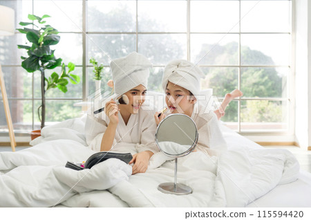 Two asian women are lying relax on a bed in white robe and towel wrapped around their head, doing makeup with a mirror and makeup zippered case. Two asian women are lying relax on a bed in white robe and towel wrapped around their head, doing makeup with a mirror and makeup zippered case. 115594420