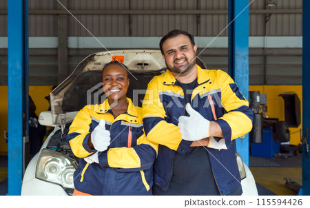 Two mechanic in work uniform and protective glove raise finger thumbs up, stand in front of a car with an open hood. Two mechanic in work uniform and protective glove raise finger thumbs up, stand in front of a car with an open hood. 115594426