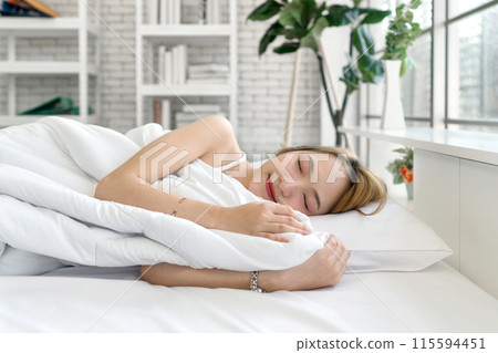 Young asian woman in white undershirt, sleep and sweet dream lying on bed in a bright, cozy room with pillow and a comforter. Side view Young asian woman in white undershirt, sleep and sweet dream lying on bed in a bright, cozy room with pillow and a comforter. Side view 115594451