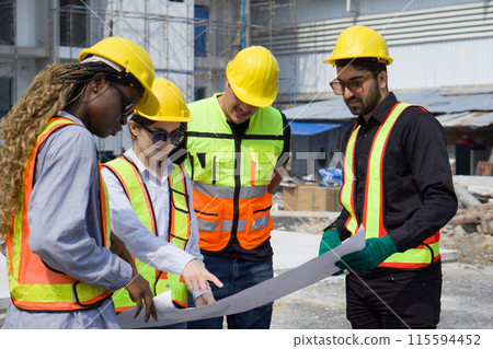 Group of construction workers gathered at a construction site reviewing some plan. Unfinished building, piles of construction material, and a partially constructed structure are in the background 115594452