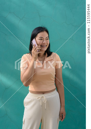 Asian woman against a turquoise wall wearing a light peach polo shirt using mobile phone Asian woman against a turquoise wall wearing a light peach polo shirt using mobile phone 115595444