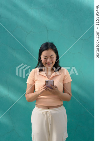 Asian woman against a turquoise wall wearing a light peach polo shirt using mobile phone Asian woman against a turquoise wall wearing a light peach polo shirt using mobile phone 115595446