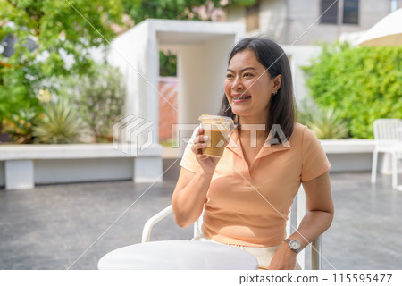 Woman sitting at an outdoor coffee shop casually dressed in an orange top and enjoys an iced coffee Woman sitting at an outdoor coffee shop casually dressed in an orange top and enjoys an iced coffee 115595477