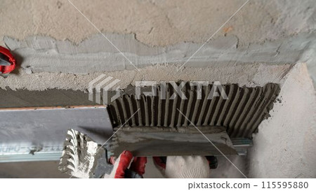 A man applying mortar to a wall with a spatula. A tiler is laying tiles in a bathroom. The technician spreads the tile adhesive with a notched trowel to adhere the tiles. A man applying mortar to a wall with a spatula. A tiler is laying tiles in a bathroom. The technician spreads the tile adhesive with a notched trowel to adhere the tiles. 115595880