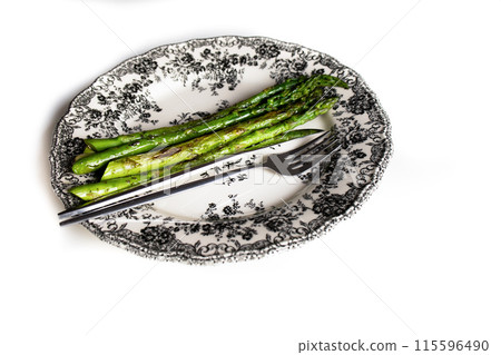 Cooked roasted asparagus and cutlery on white plate with black floral print Cooked roasted asparagus and cutlery on white plate with black floral print 115596490