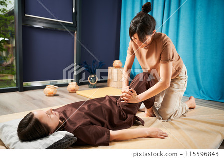 Masseuse making thai yoga massage. Woman therapist doing traditional massage treatment for female patient indoors. Client lying on mat, meditating, enjoying hand massage in calm atmosphere. Masseuse making thai yoga massage. Woman therapist doing traditional massage treatment for female patient indoors. Client lying on mat, meditating, enjoying hand massage in calm atmosphere. 115596843