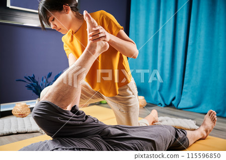 Masseuse making thai yoga massage. Woman therapist doing traditional massage treatment, stretching male patient's body. Client lying on mat, enjoying hand massage in calm atmosphere. 115596850