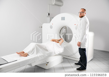 Medical computed tomography or MRI scanner. Male radiologist presses MRI button to examine female patient, looking to the camera. Concept of healthcare and modern diagnostics. 115596859