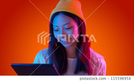 Internet surfing. Reading tablet. Hipster woman using modern device typing social media communication watching video blue neon light isolated on red gradient empty space background. 115596958