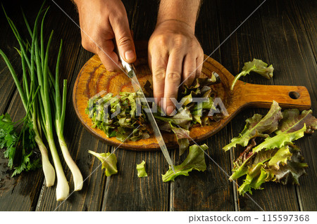 Chef preparing salad of cos lettuce and onions with hands and knife on kitchen table. Slicing romaine violets on wooden board for vegetarian food 115597368