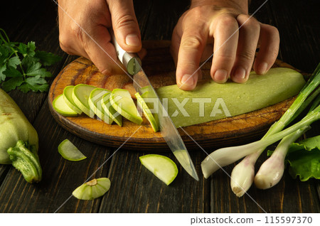 Cooking vegetable marrow with vegetables by the chef hands according to a special recipe in the restaurant kitchen. Slicing zucchini with a knife Cooking vegetable marrow with vegetables by the chef hands according to a special recipe in the restaurant kitchen. Slicing zucchini with a knife 115597370