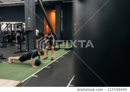 Remote view of beginner sportsman exercising with heavy medicine ball while personal trainer encouraging during cross training in health club. Fitness guy doing weight exercises with coach assistance 115599440