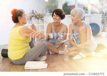 Mature women talking to each other sitting on the floor in fitness studio Mature women talking to each other sitting on the floor in fitness studio 115603771