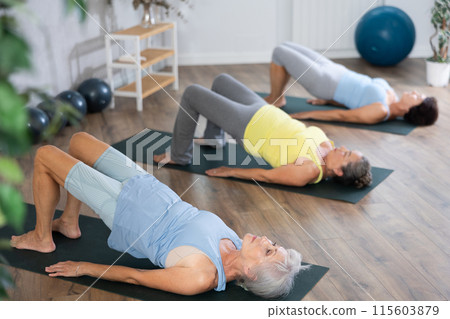 Elderly woman in company of hatha yoga lovers performs bridge pose, setu bandha sarvangasana 115603879