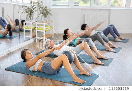 Women maintaining active lifestyle exercising with ball sitting on yoga mat during group workout in fitness 115603919
