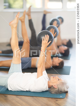 Senior woman performing leg lifts with exercise ball in group Pilates class 115603964