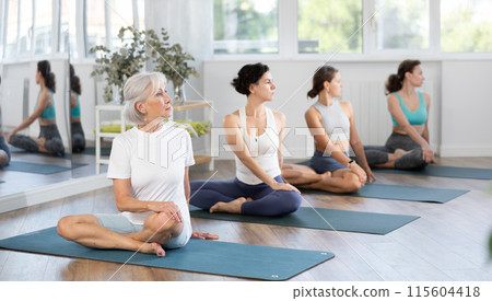 Senior woman sitting in lotus pose with side twist at yoga class Senior woman sitting in lotus pose with side twist at yoga class 115604418