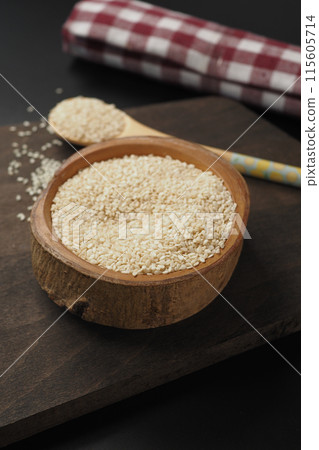 A wooden bowl filled with white sesame seeds, accompanied by a spoon, set on a dark surface A wooden bowl filled with white sesame seeds, accompanied by a spoon, set on a dark surface 115605714