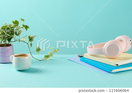 Office theme photo from front angle standing out blue background, a pile of notebook displayed at the right side with a headphone placed on. A coffee cup and plant pots on placed on left for decorate Office theme photo from front angle standing out blue background, a pile of notebook displayed at the right side with a headphone placed on. A coffee cup and plant pots on placed on left for decorate 115606064