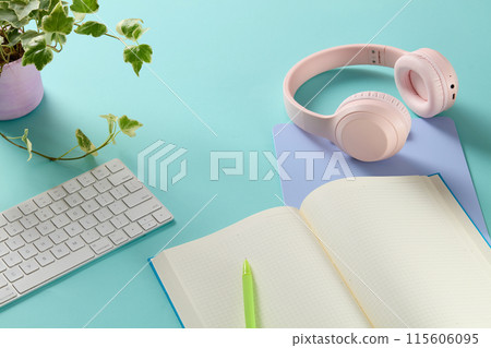 The blue working desk surface features a open white notebook, next to a pink headphone, a white keyboard and a pot of green plant. High angle shot photo with vacant space for product presentation The blue working desk surface features a open white notebook, next to a pink headphone, a white keyboard and a pot of green plant. High angle shot photo with vacant space for product presentation 115606095