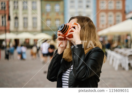 Woman taking picture using orange instant camera. Tourist captures memories during travel with vintage camera. Female walking around city street 115609031