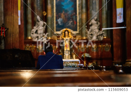 Back view of man in blue jacket prays in catholic church. He is seated in pew with his head bowed in prayer. Altar is visible in background. 115609053