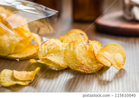Crispy Potato Chips Spilled From  Plastic Bag on a Wooden Table 115610840