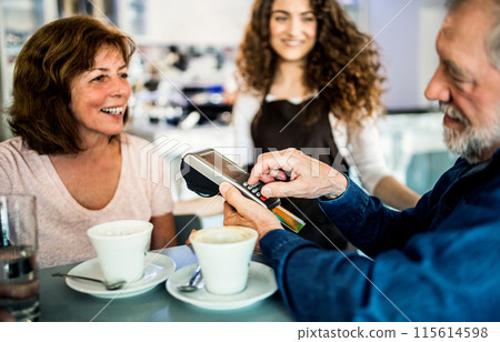 Elderly couple paying for their coffe with debit card. University student working part-time in cafe. 115614598