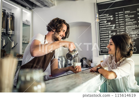 Handsome barista working in coffee shop, preparing coffee for female customer at counter. University student working part-time in cafe. Handsome barista working in coffee shop, preparing coffee for female customer at counter. University student working part-time in cafe. 115614619