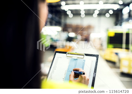 Manager woman in reflective safety clothes, holding clipboard with documents, controlling goods in warehouse. 115614720
