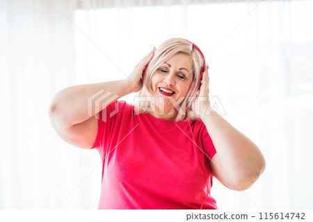 Portrait of beautiful young overweight woman at home, listening music via headphones, having fun. 115614742