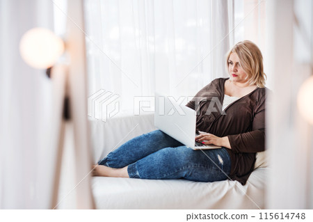 Overweight woman working on laptop from homeoffice, sitting on sofa with legs up. 115614748