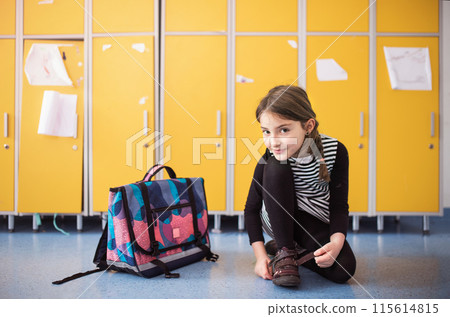Portrait of schoolgirl, putting shoes on by lockers. Student girl with bag in hallway, after last day in elementary school before summer break. 115614815