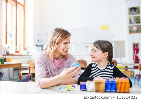 Hardworking teacher learning with young schoolgirl in classroom, explaining her shapes and math, geometry. 115614822