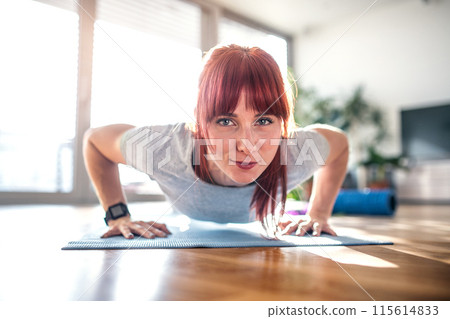 Beautiful redhead woman's home fitness routine, doing pushups. Home workout on gym mt in living room, sporty woman wearing sportswear. 115614833