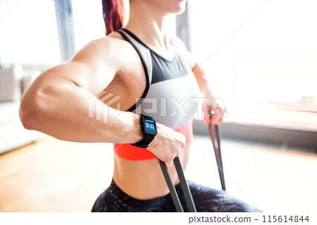 Beautiful redhead woman's home fitness routine with resistance band. Home workout on gym mat in living room, sporty woman wearing sportswear. 115614844