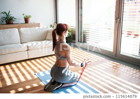 Beautiful redhead woman's home fitness routine with resistance band. Home workout on gym mat in living room, sporty woman wearing sportswear. 115614879