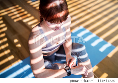 Beautiful redhead woman checking her performance on smartwatch after home fitness routine. Home workout on gym mt in living room, sporty woman wearing sportswear. Beautiful redhead woman checking her performance on smartwatch after home fitness routine. Home workout on gym mt in living room, sporty woman wearing sportswear. 115614880