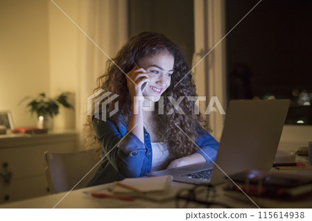 Beautiful university student is writing her thesis on a laptop at night, sitting at home at table, phone calling on smartphone. Preparing for final exam, studying. 115614938
