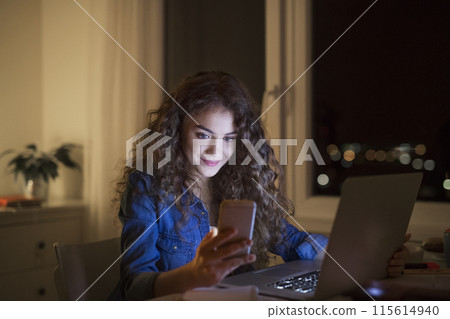 Beautiful university student is writing her thesis on a laptop at night, sitting at home at table, scrolling on smartphone. Preparing for final exam, studying. 115614940