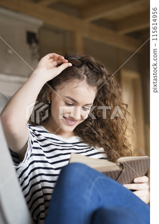 Female student sitting in armchair, reading book, studying for final exam at university. Beautiful woman relaxing at home. Female student sitting in armchair, reading book, studying for final exam at university. Beautiful woman relaxing at home. 115614956